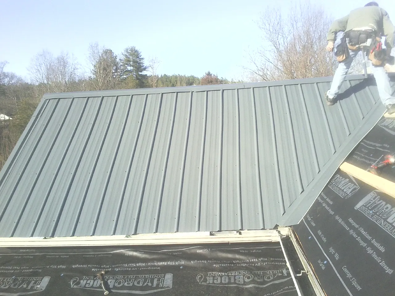 Crew installing dark green standing seam panels over synthetic underlayment in Spring Lake